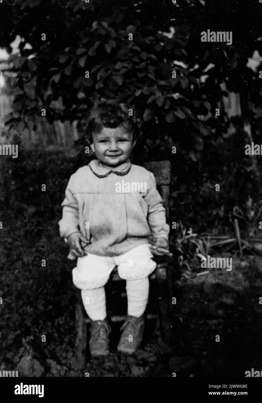 Schwarz-Weiß-Porträt eines 3 Jahre alten lächelnden Mädchens, das auf einem Stuhl in ländlicher Umgebung sitzt, Ende 60s, Bulgarien, Europa, Balkan Stockfoto