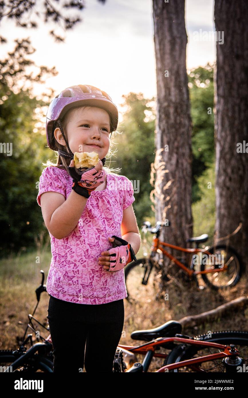 Ein kleines Mädchen mit einem Fahrradhelm, das vor dem Fahrrad steht, isst einen Apfel Stockfoto