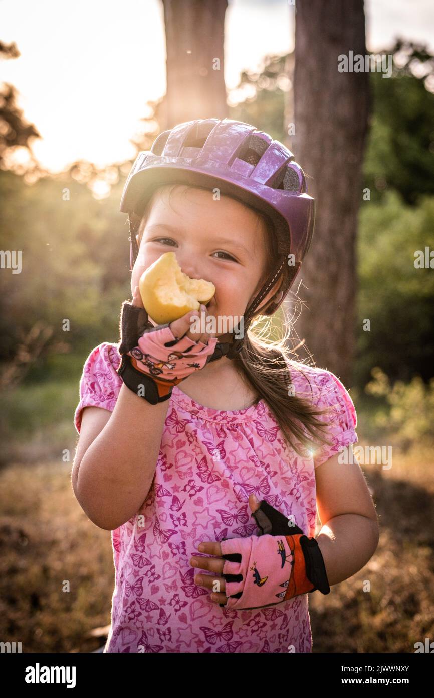 Ein kleines Mädchen mit Fahrradhelm frisst einen Apfel Stockfoto