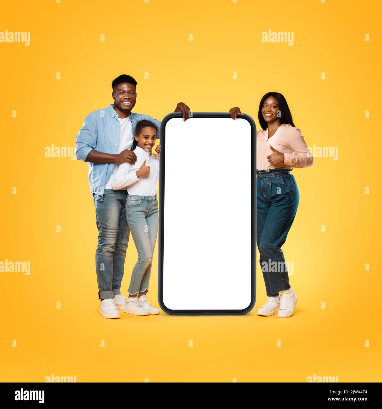 Glückliche schwarze Familie, die in der Nähe des riesigen Handys mit weißem, leeren Bildschirm steht, Daumen hoch gestikuliert und lächelt Stockfoto