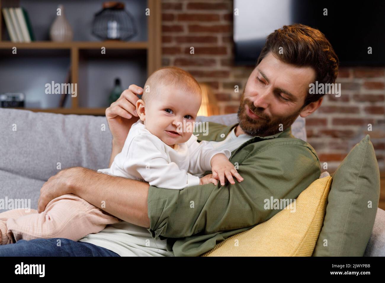 Papa spielt mit seinem kleinen Baby auf der Couch in einer gemütlichen Wohnung. Väterliche Liebe. Vater umarmt und küsst seine kleine Tochter. Stockfoto