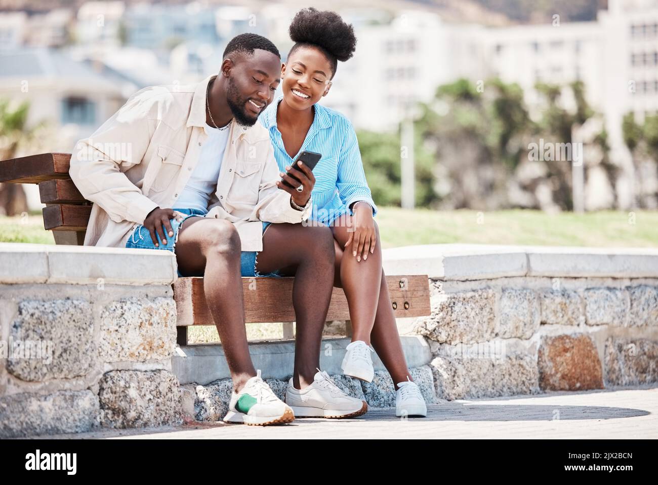 Schwarzes Paar, Liebe und Telefon beim Surfen in sozialen Medien, im Internet oder beim Ansehen eines Videos, während Sie auf einer Parkbank sitzen. Glücklicher Mann und Frau auf einem Stockfoto