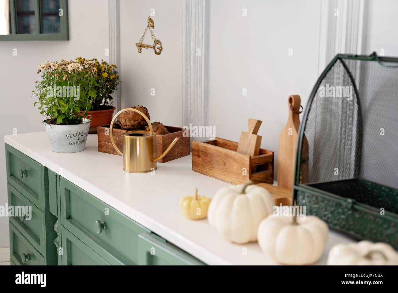 Küche mit grünen Vintage-Möbeln, Pendelleuchten, Marmor-Arbeitsplatte mit Blumen und Kürbissen, Töpfe mit Herbstblumen, Schrank mit verschiedenen Tassen Stockfoto