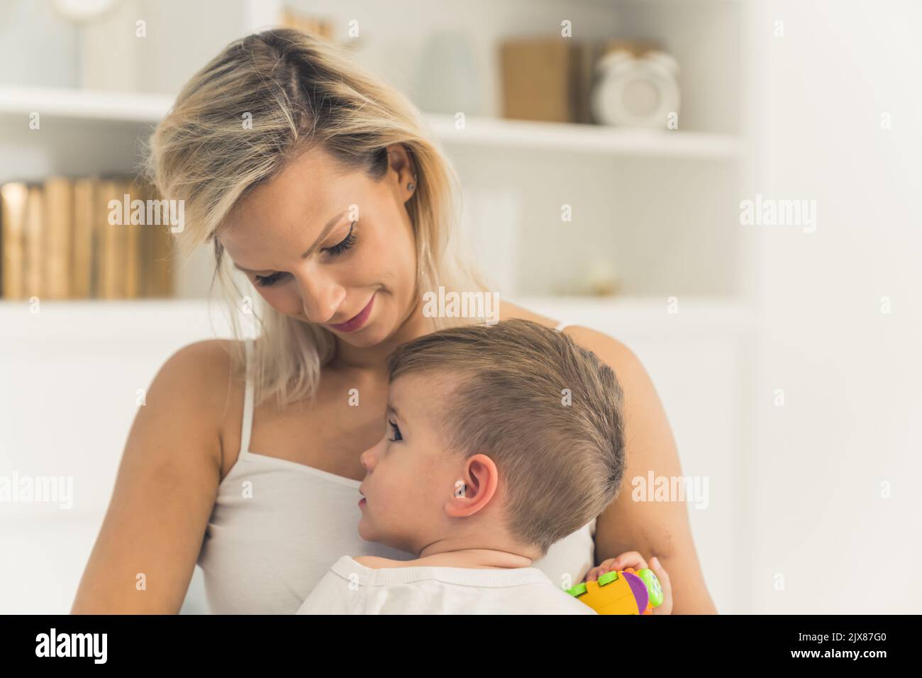 Nahaufnahme Porträt präsentiert wirklich schöne Bindung zwischen Eltern und kleinen Kind. Kaukasischer braunhaariger Kleinkind, der auf dem Schoß seiner Mutter sitzt. Stolze glückliche Mutter im weißen Tank Top, die ihr Kind ansieht. Hochwertige Fotos Stockfoto