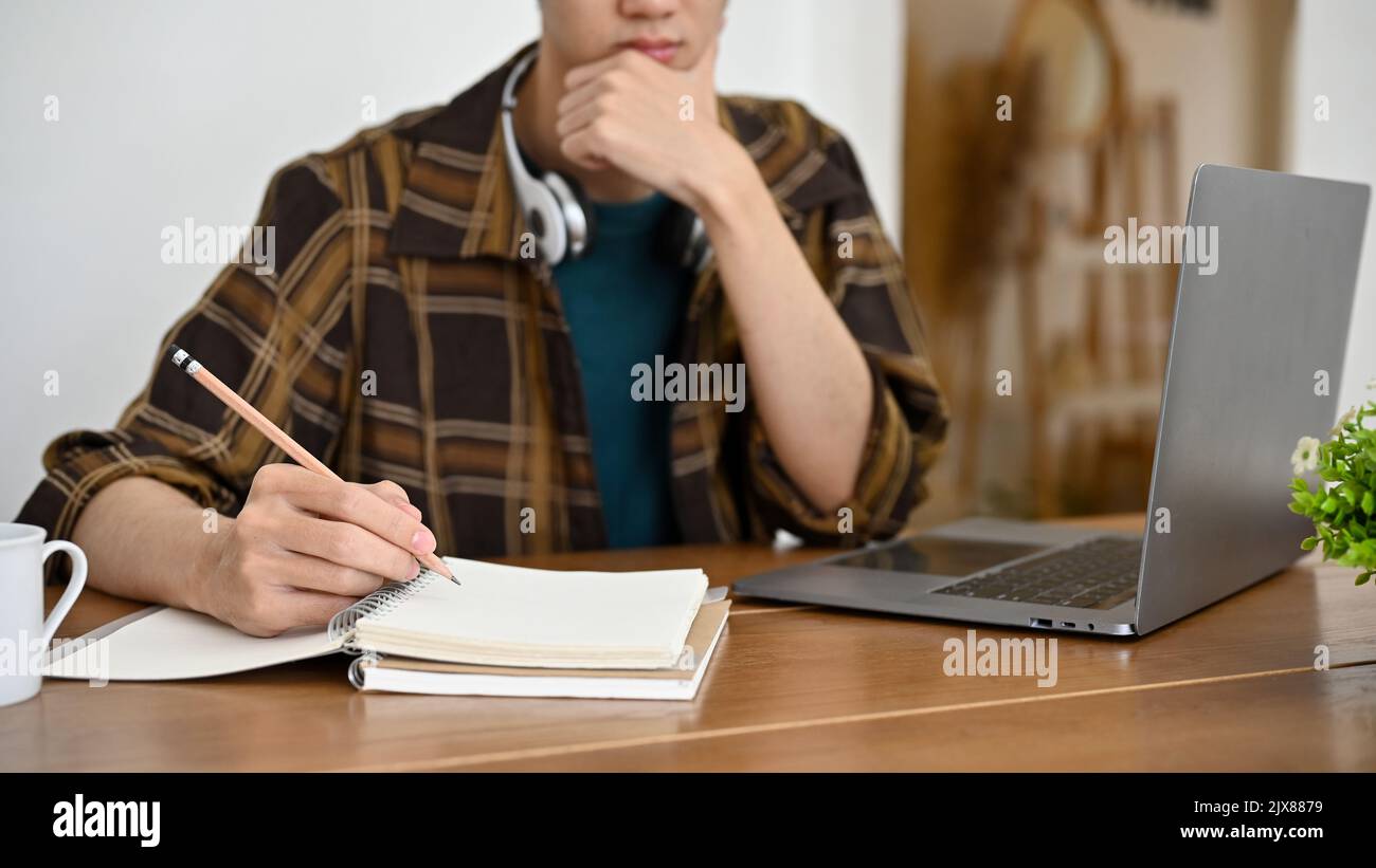 Nachdenklicher junger asiatischer Freiberufler oder Student, der an seinem Schreibtisch arbeitet, pensiver denkt, sein Projekt zu planen, Hausaufgaben macht. Zugeschnittenes Bild Stockfoto