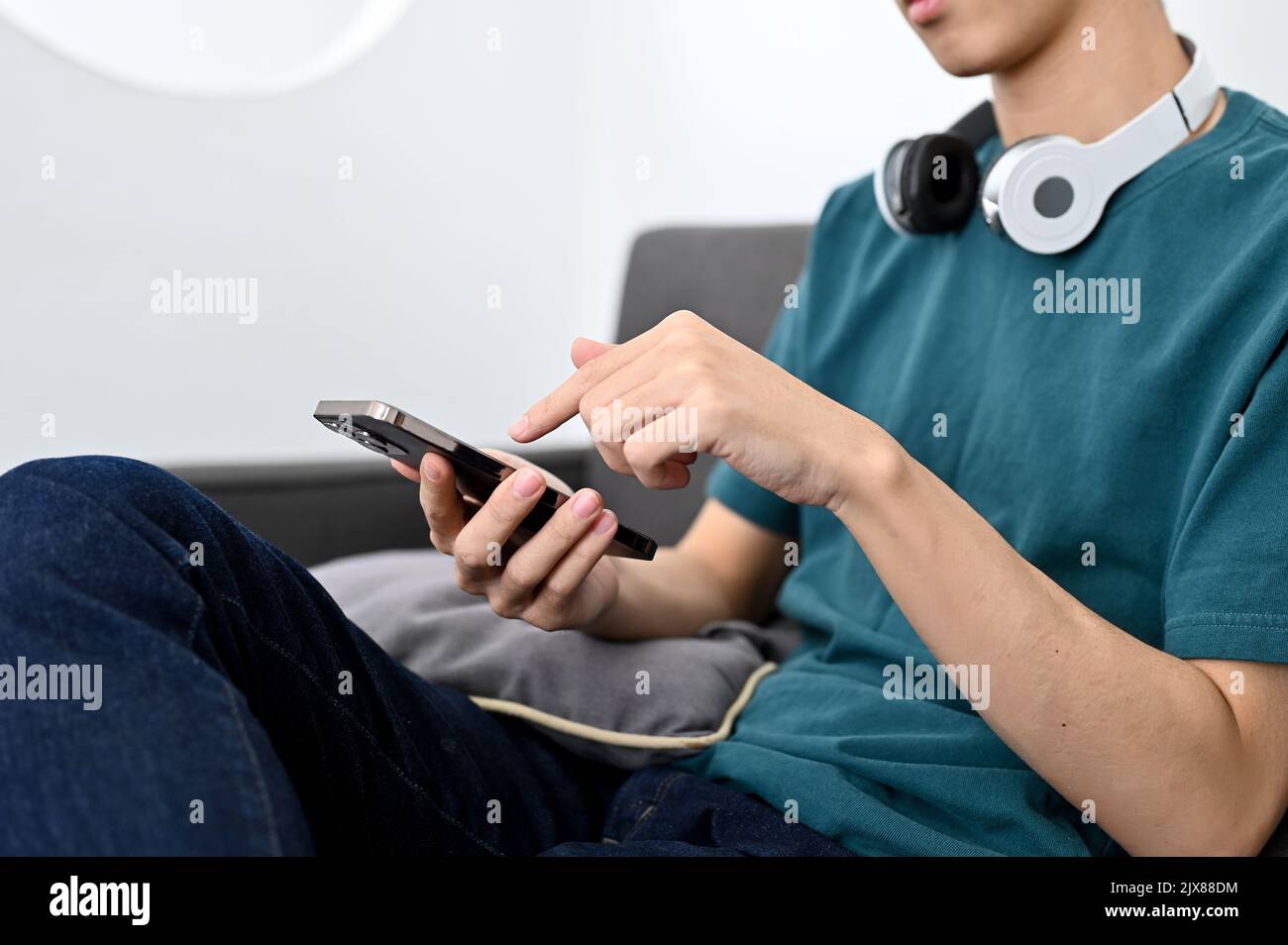 Ein tausendjähriger asiatischer Mann, der auf dem Sofa sitzt und sein Smartphone benutzt. Stockfoto