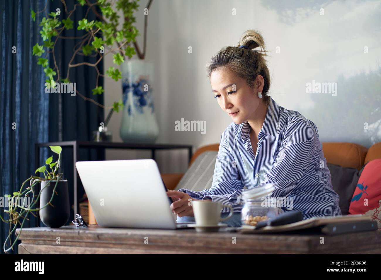 Reife professionelle asiatische Frau sitzt auf der Couch arbeiten von zu Hause aus mit Handy und Notebook-Computer Stockfoto