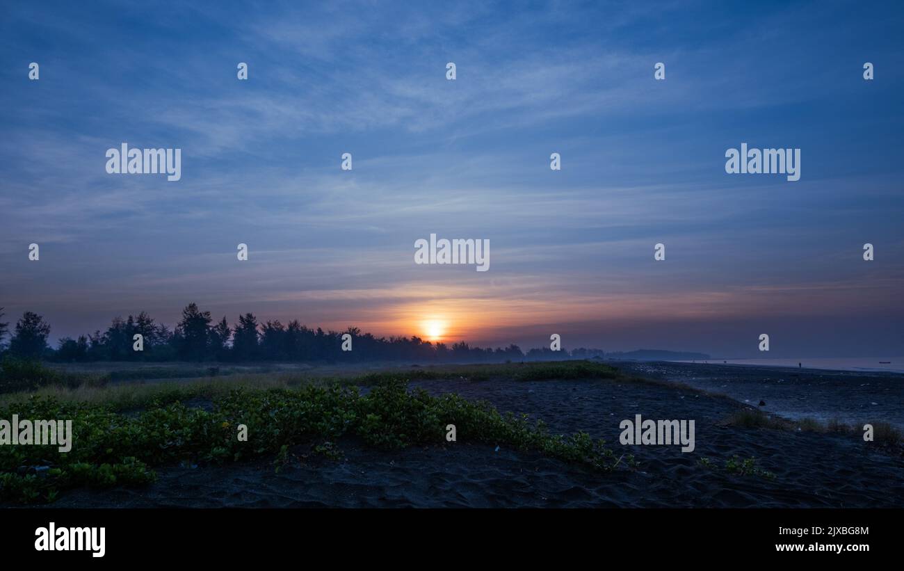Die goldorange Sonne geht durch die Bäume hinter dem Busch am Strand von Suruchi in Vasai auf und durchbricht die Wolkenlinien des blauen Himmels. Stockfoto