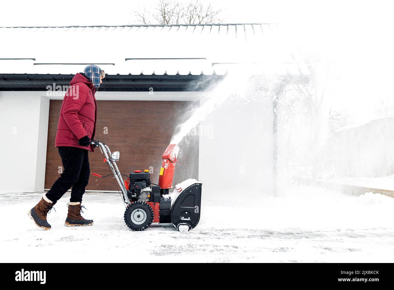 Schneegebläse mit Benzin in Aktion. Mann draußen vor dem Haus mit Schneefräse Maschine. Schneeräumung, Werferassistent im Winter außerhalb des Hauses. Junger Arbeiter, der während des Schneesturms Schnee bläst Stockfoto