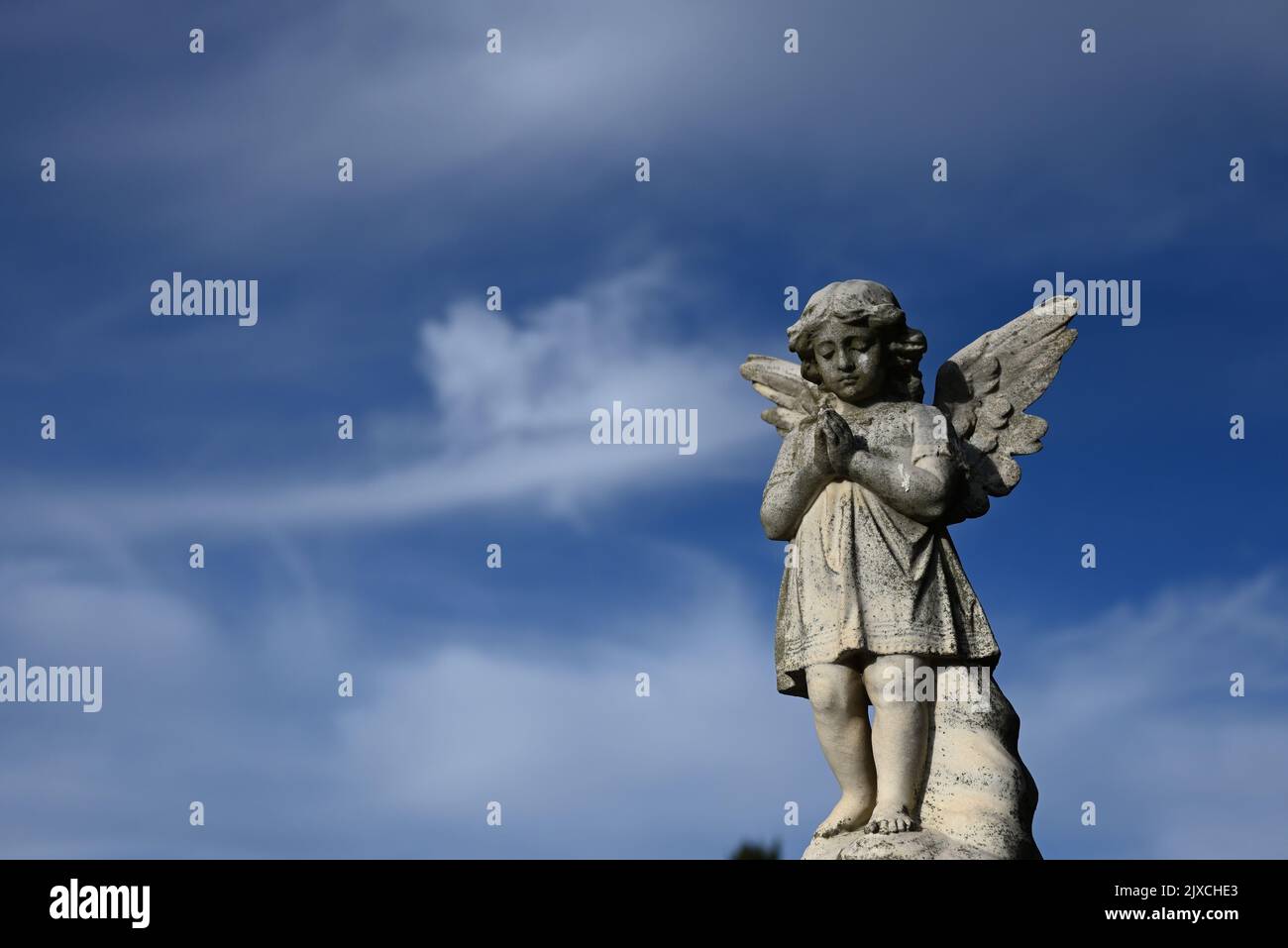 Steinskulptur eines Kinderengels oder Cherubs mit gebetteten Händen, mit blauem Himmel und flauschigen weißen Wolken im Hintergrund Stockfoto