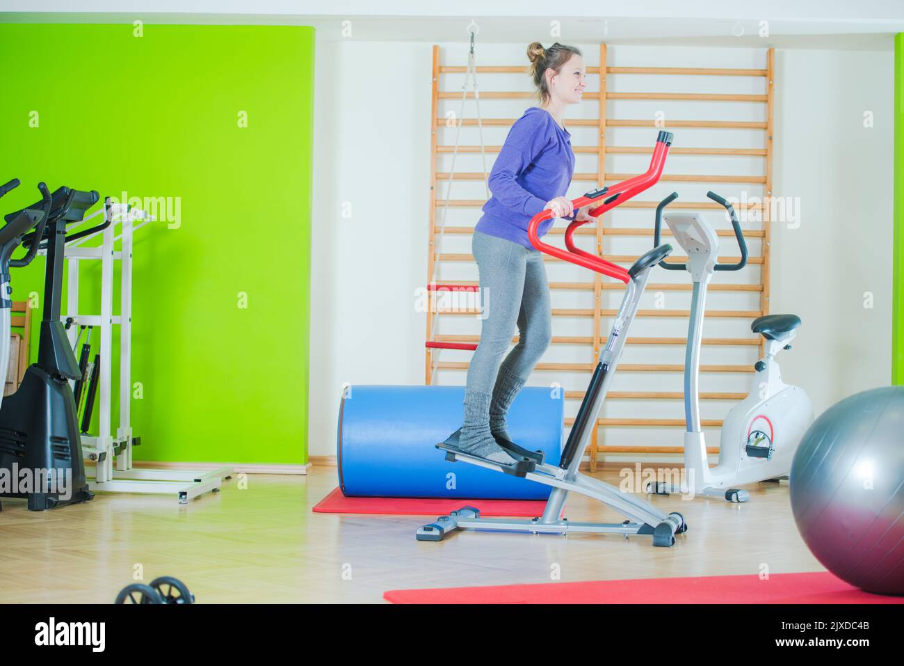 Lächelnde junge Frau, die ihr Fitness-Workout auf einem Cross-Trainer-Gerät macht. Fitnessraum mit verschiedenen Trainingsgeräten im Hintergrund. Stockfoto
