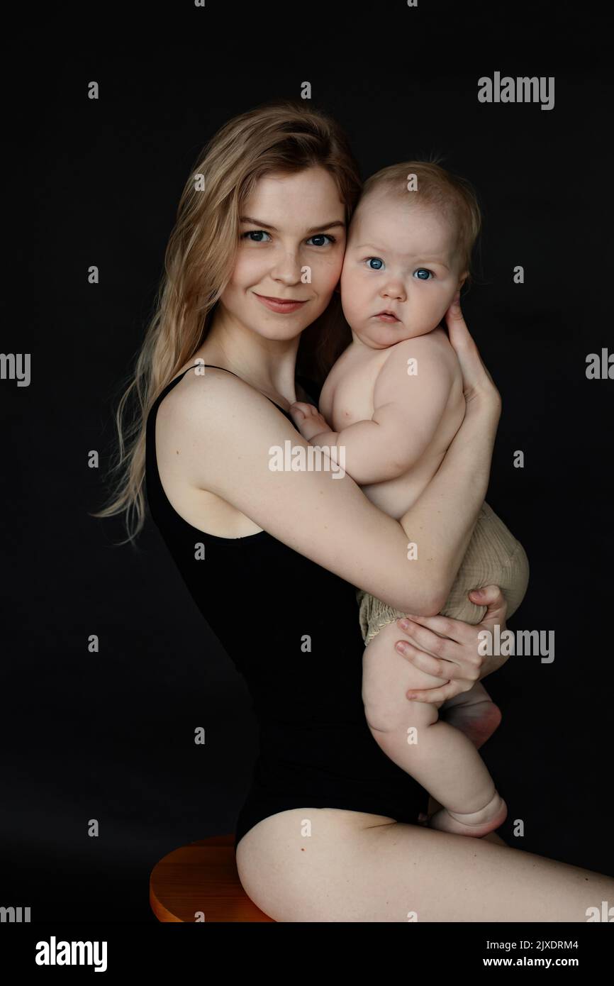 Vertikales Porträt der frohen Mutter umarmt kleines Baby in den Armen, Blick auf die Kamera auf schwarzem Hintergrund. Kinder lieben und kümmern sich um sie Stockfoto
