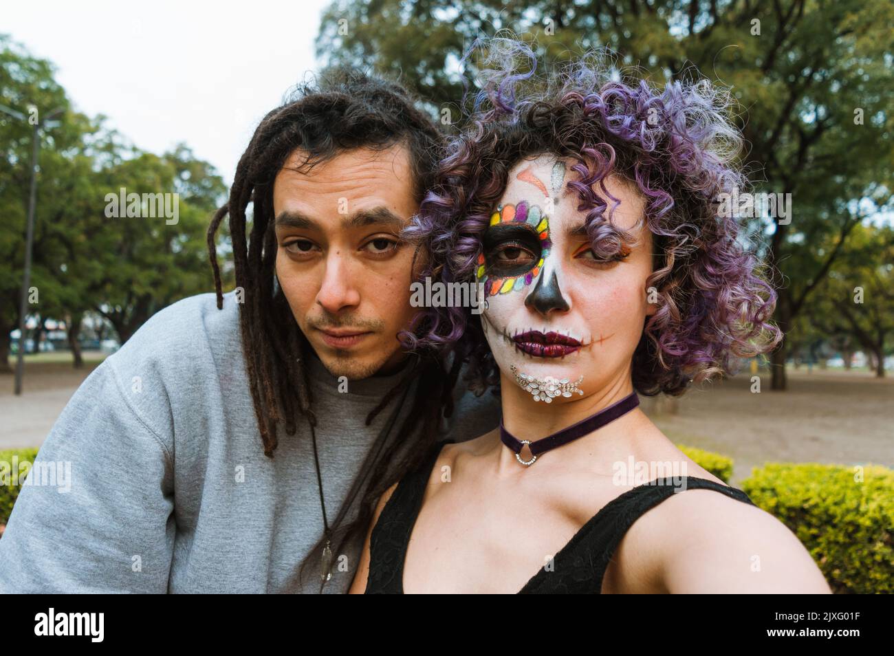 Porträt eines jungen lateiners und einer jungen Frau im Freien, die einen Tag des toten Selfies mit einem bemalten catrina-Gesicht in einem Park mit einem Lächeln und Blick auf die Kamera machen Stockfoto