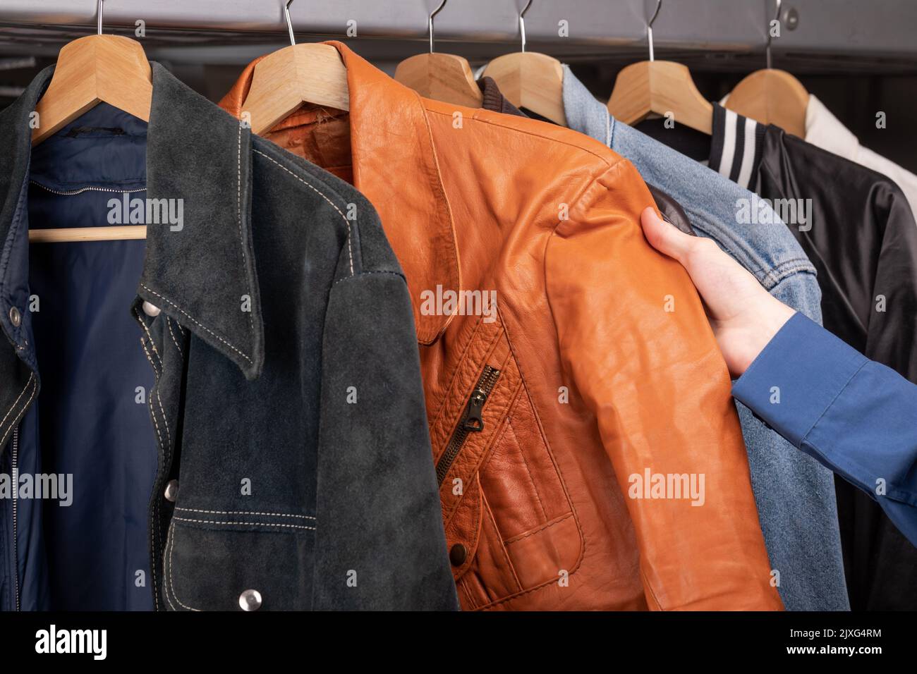 Der Mann überprüft die orangefarbene Lederjacke in einem Second-Hand-Geschäft. Verschiedene Vintage-Jacken hängen an Kleiderständer. Thrifting und Nachhaltigkeit in der Bekleidungskonce Stockfoto