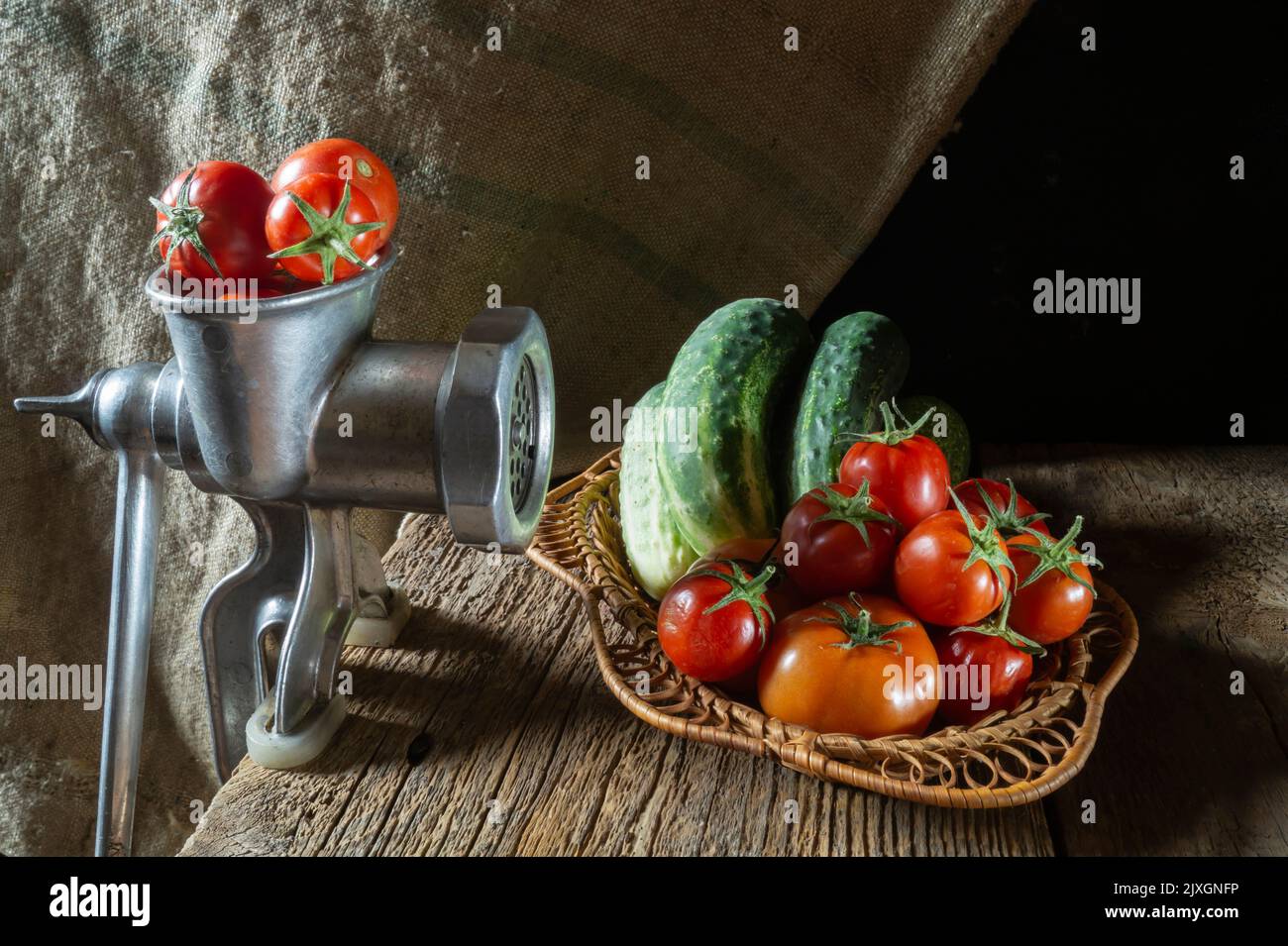 Reife Tomaten und Gurken in einem Korb auf einem Holztisch. Fleischwolf mit Tomaten auf dem Hintergrund von Sackleinen Stockfoto