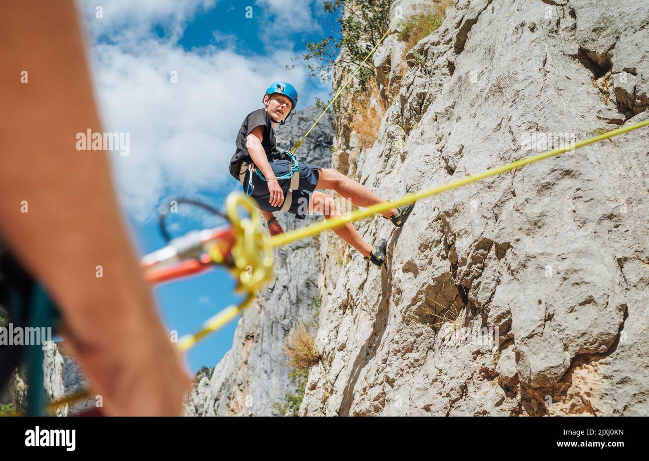Teenager Junge auf der natürlichen Klippe Kletterwand. Der Junge hing mit einem Klettergurt an einem Seil und der Vater brachte ihn mit einem bela auf den Boden Stockfoto