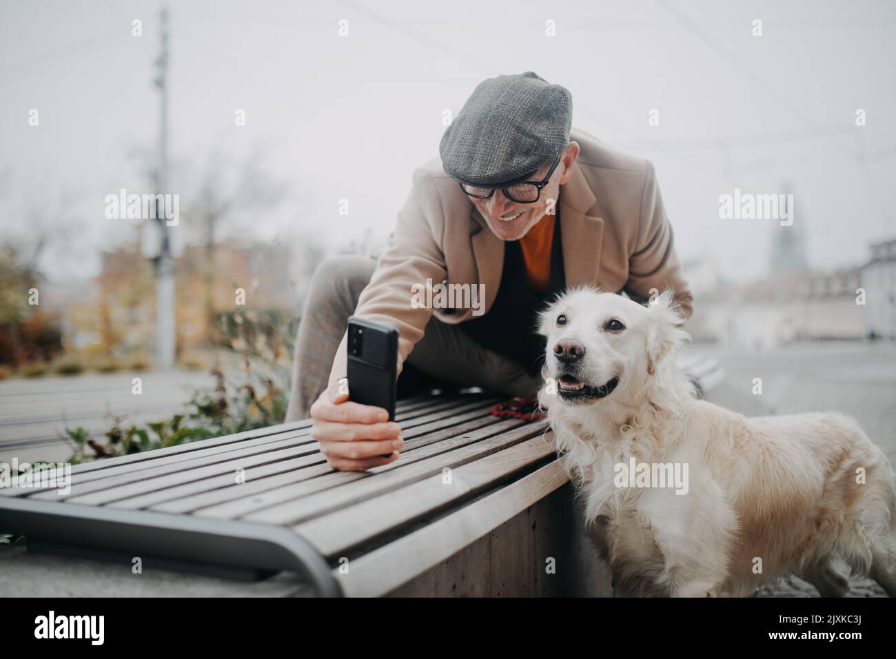Eleganter älterer Mann mit Smartphone, der Selfie mit seinem Hund nimmt und in der Winterstadt im Freien läuft. Stockfoto
