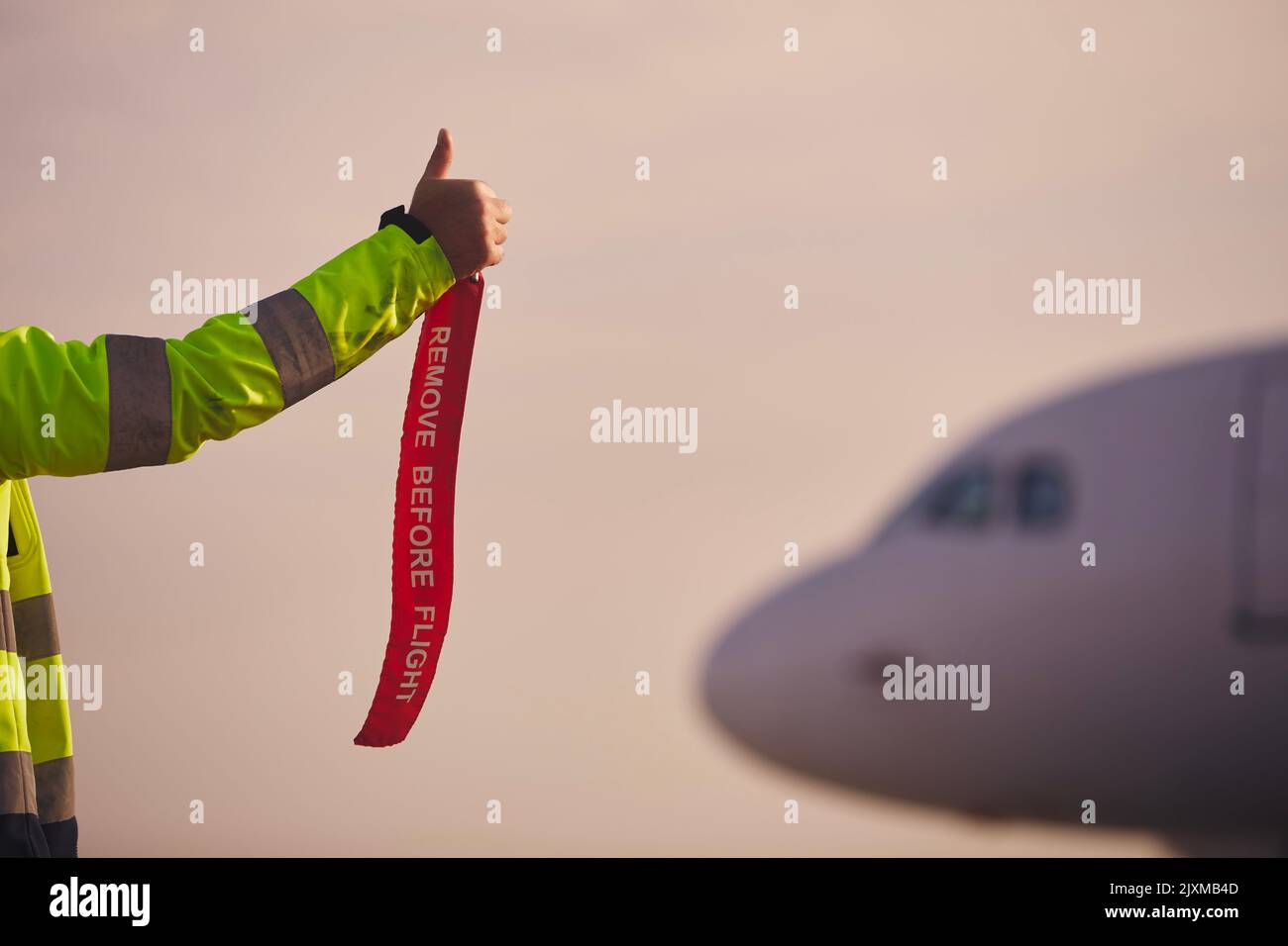 Mitglied der Bodenbesatzung mit der roten Flagge vor dem Flug entfernen und Daumen hoch zum Flugzeug zeigen. Verkehr am Flughafen bei Sonnenuntergang. Stockfoto