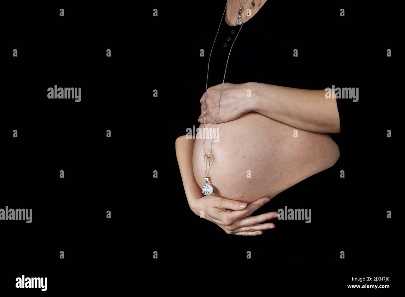 Detail des Bauches einer schwangeren Frau mit einer kleinen Glocke am Bauchnabel, um das Baby ruhig zu halten. Alles schwarz, Kleidung und Hintergrund, wir sehen nur ihre Hände Stockfoto