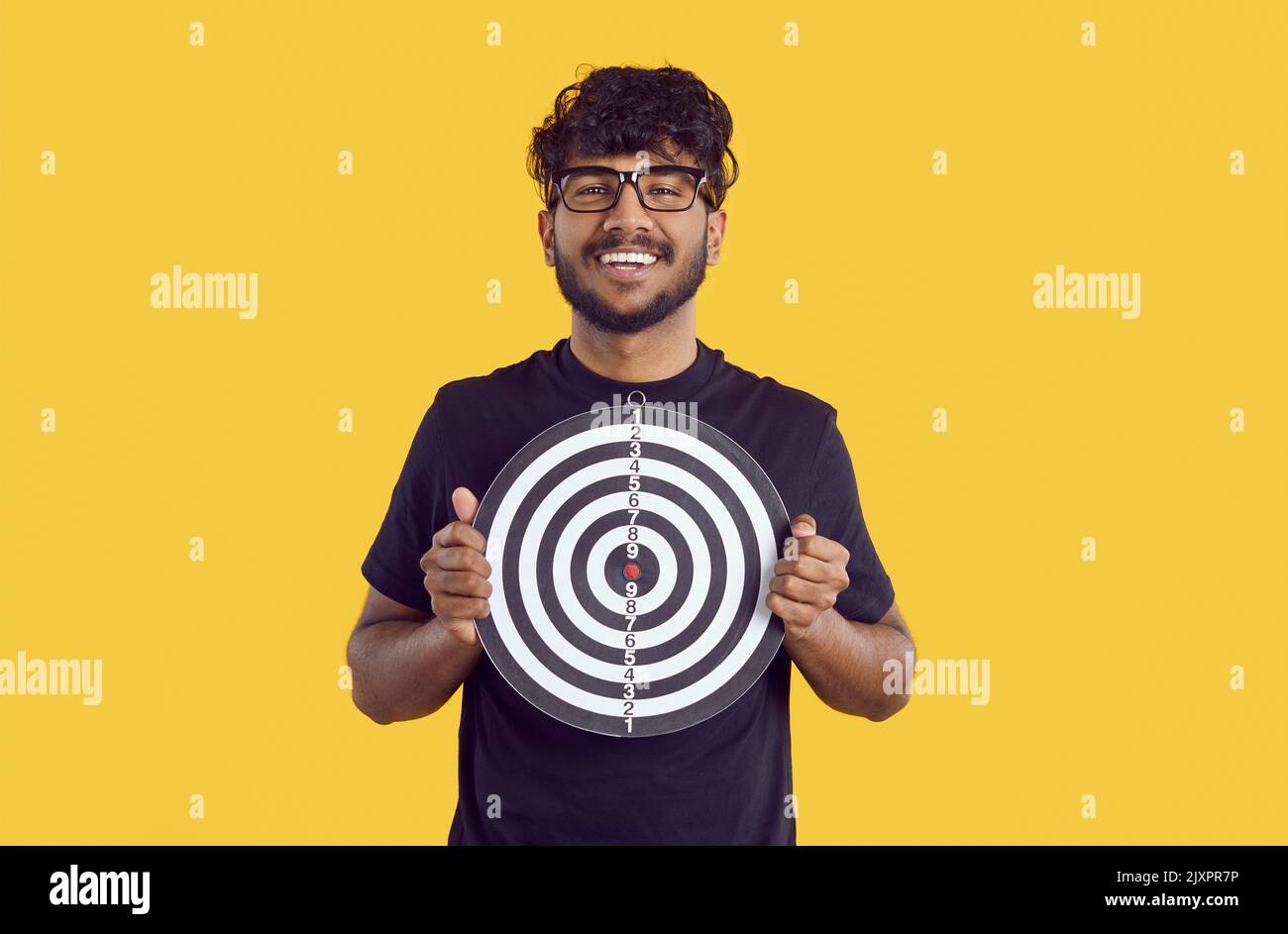 Portrait von indischen Kerl hält Darts Board mit glücklich und selbstbewusst Gesicht auf orangenen Hintergrund. Stockfoto