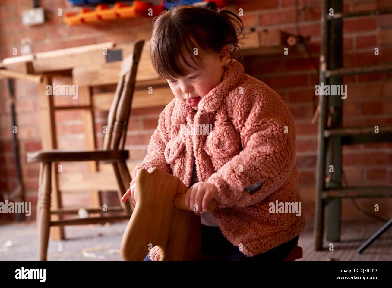 Mädchen Mit Down-Syndrom Sitzen Und Spielen Auf Hölzernen Schaukelpferd In Garage Workshop Zu Hause Stockfoto