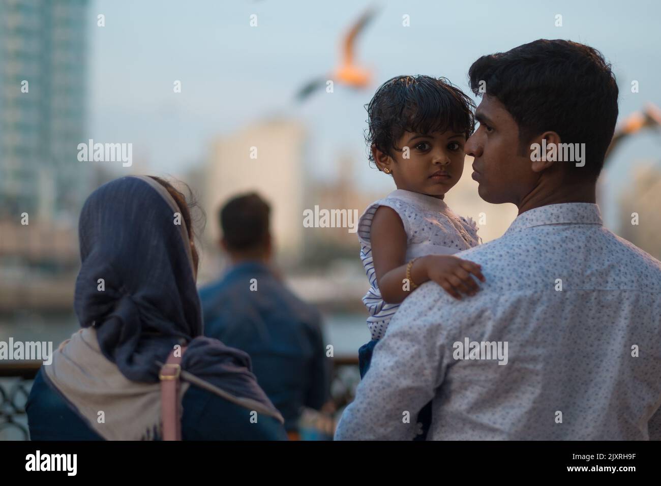 Vater trägt eine junge Tochter auf seinem linken Arm. Mädchen schaut in die Kamera, während der indische Mann Gesicht im Profil hat. Mutter, Frau mit Schleier, steht neben ihnen. Stockfoto