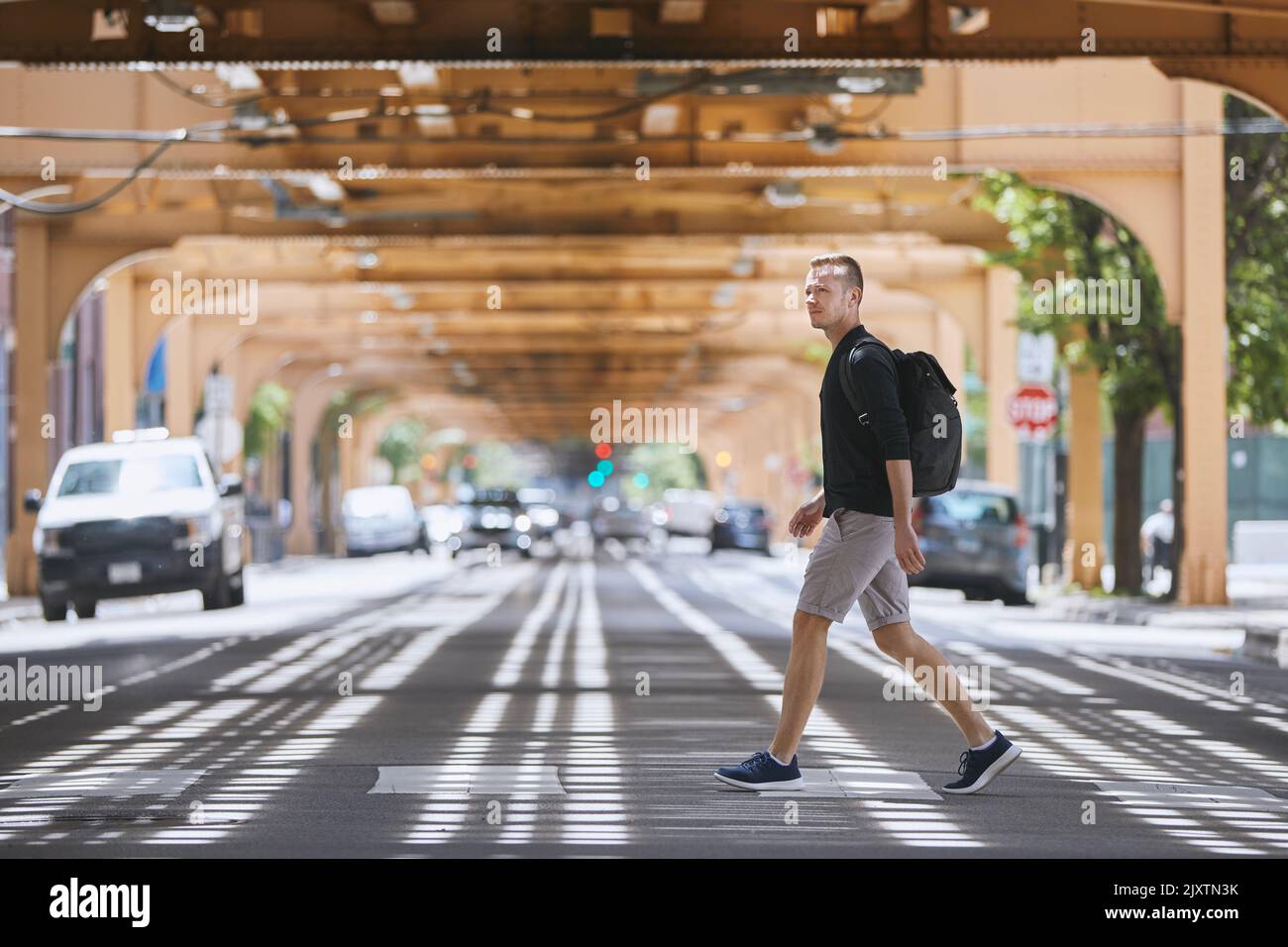 Seitenansicht eines erwachsenen Mannes mit Rucksack, der auf einem Fußgängerüberweg über die Hochbahn der öffentlichen Verkehrsmittel läuft. Chicago, Usa Stockfoto