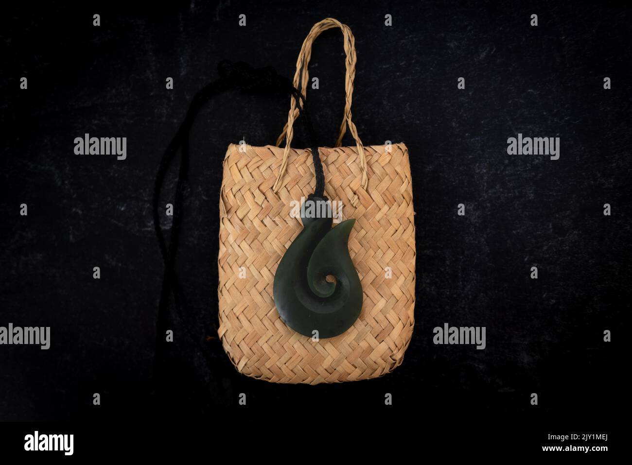Pounamu Fischhaken Halskette auf gewebten Tasche auf dunklem Hintergrund. Aotearoa, Neuseeland, Maori. Stockfoto