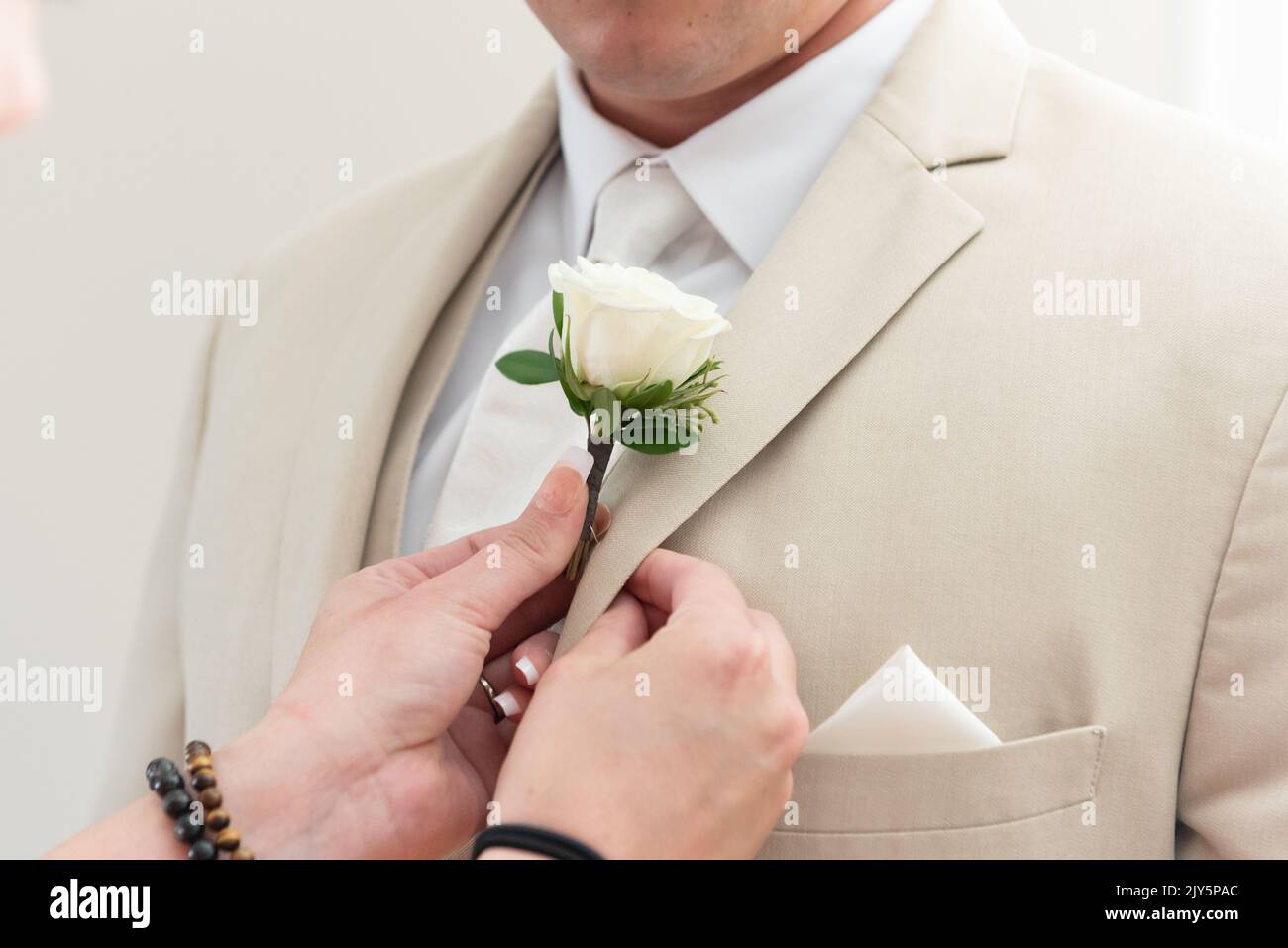 Der Bräutigam hat helfende Hände mit einer Maniküre, die ihm dabei hilft, seine Blumenrosenboutonniere auf das Revers seines Anzugs zu pinnen. Stockfoto