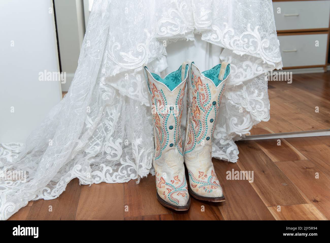 Weißes Spitzenkleid der Braut wird für eine westliche Hochzeitszeremonie mit weißen bestickten Cowgirl-Stiefeln kombiniert. Stockfoto