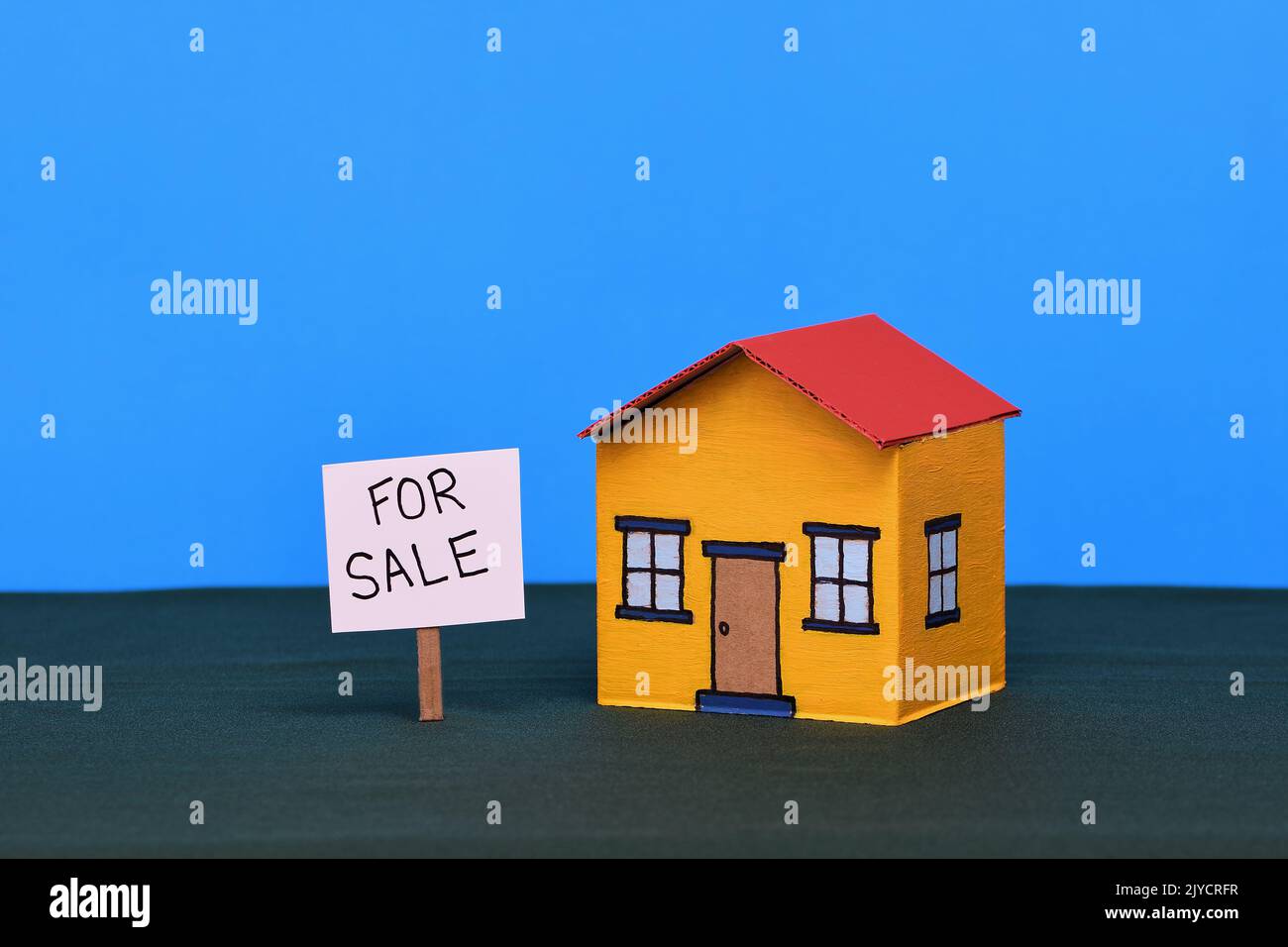 Ein gelbes und rotes Spielzeugkartonhaus in der Mitte des Rahmens auf einem dunkelgrünen Sockel und blauem Hintergrund in sanfter Beleuchtung mit einem Schild „Realestate for Sale“ Stockfoto