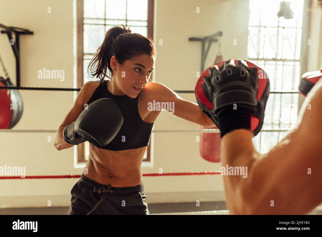 Hübsche junge Frau trainiert im Boxring mit Partner in der Nähe von rotem Boxsack und anderen Sparringausrüstungen Stockfoto