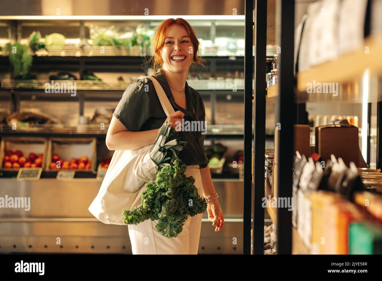 Eine Kundin lächelt fröhlich, während sie eine Tasche mit frischem Bio-Gemüse in einem Lebensmittelgeschäft trägt. Glückliche junge Frau Lebensmitteleinkauf in einem Tren Stockfoto