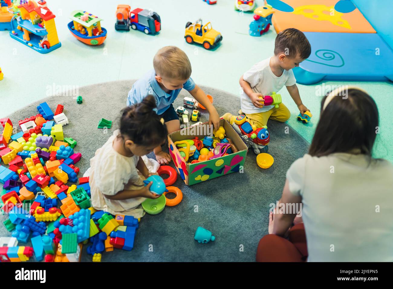 Kleinkinder und ihre Kindergärtnerin spielen mit Plastikbausteinen und farbenfrohen Autospielzeugen in einem Kinderzimmer. Frühe Entwicklung von Gehirn und Fähigkeiten. Hochwertige Fotos Stockfoto