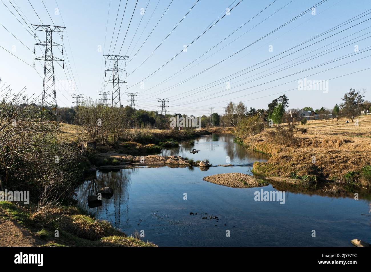 Elektrische Übertragung Oberleitung Stromleitungen, die über einen idyllischen Bach in einer städtischen Grünfläche verlaufen Stockfoto