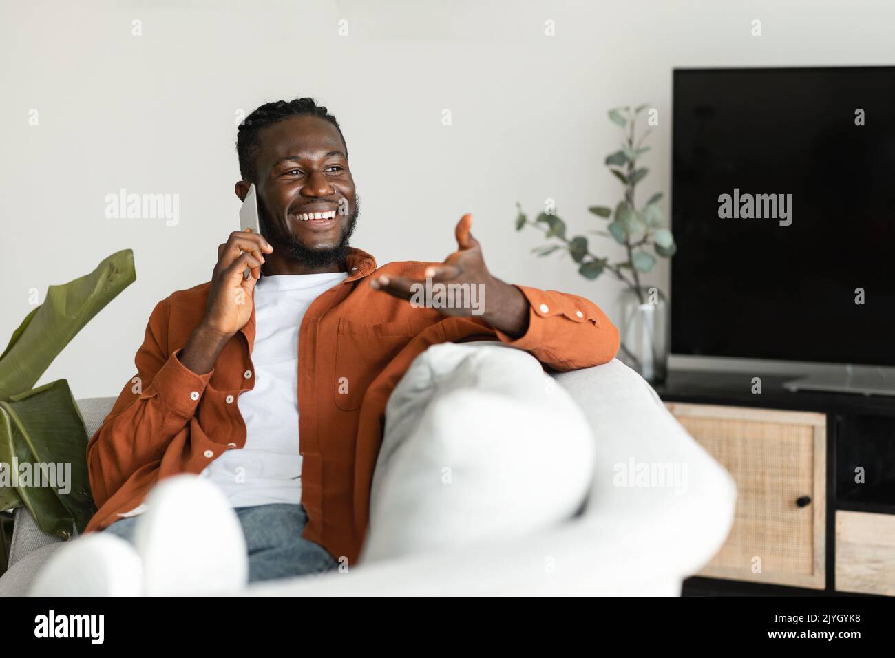 Anruf tätigen. Glücklicher afroamerikanischer Mann, der auf dem Smartphone spricht und gestikiert, sich zu Hause auf dem Sofa ausruht, freien Platz Stockfoto