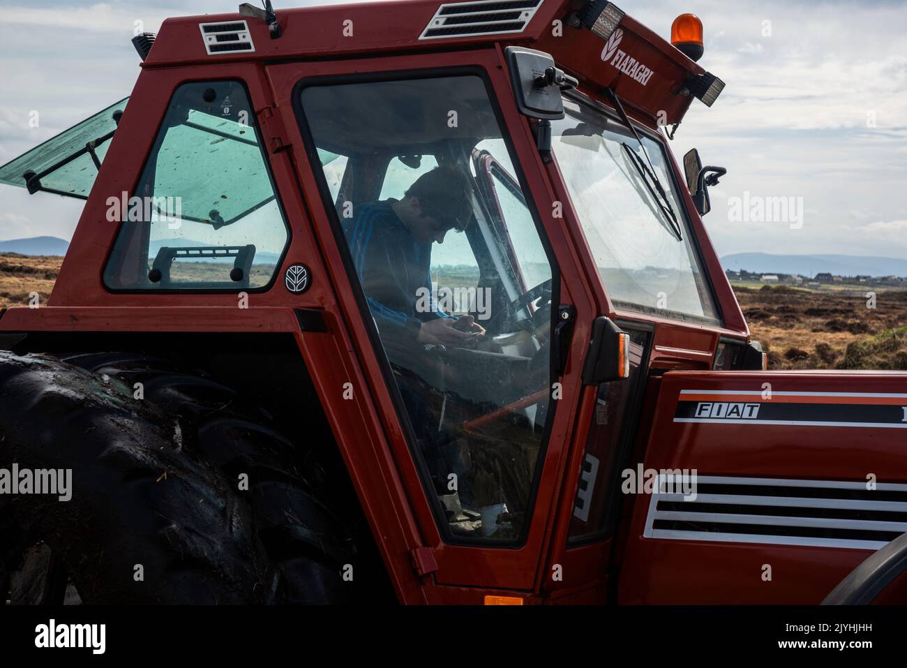 Belmullet, County Mayo, Irland, 4-9-2020. Ein Junge in einem Traktor mit einem Trichter dahinter. Er überprüft sein Telefon, während der Trichter wieder voll wird. Stockfoto