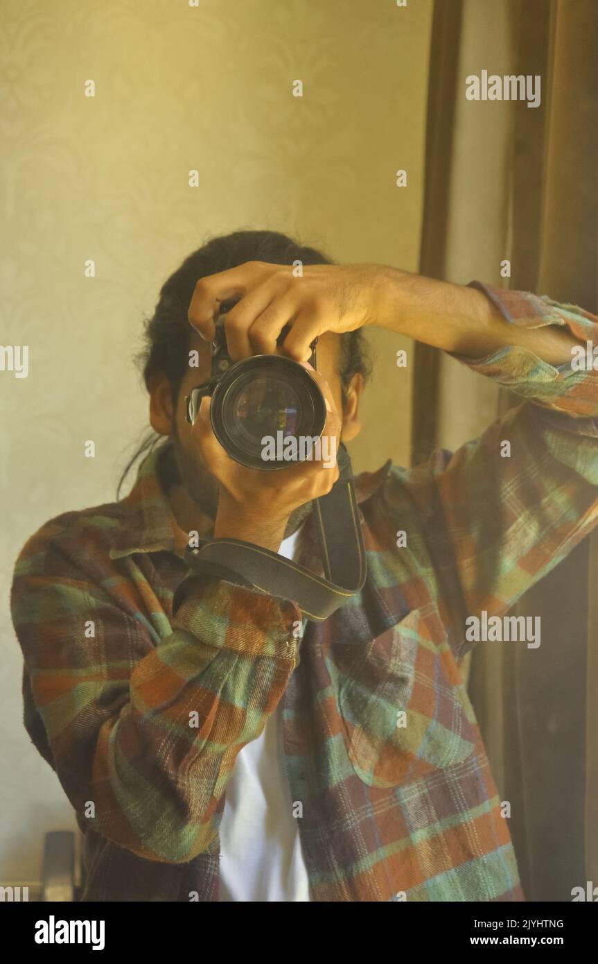 Die Taille eines männlichen Fotografen, der durch den Sucher seiner DSLR-Kamera schaut und vor dem Spiegel im Raum fotografiert. Stockfoto