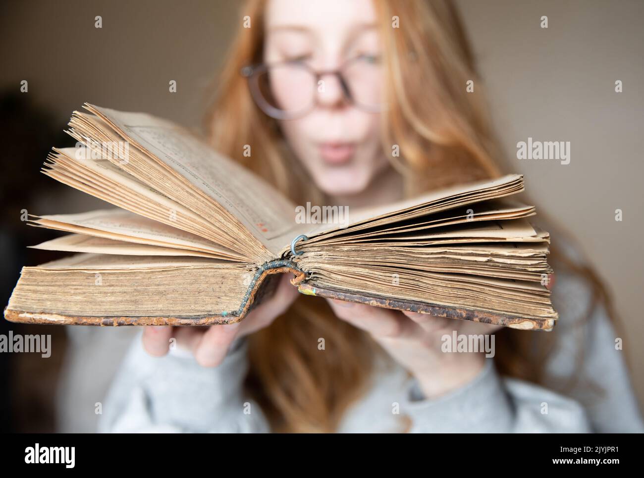 Ein offenes Vintage-Buch in den Händen eines niedlichen Teenagers mit Brille und roten Haaren, in einem halbdunklen Raum, der vom Licht des Fensters beleuchtet wird Stockfoto