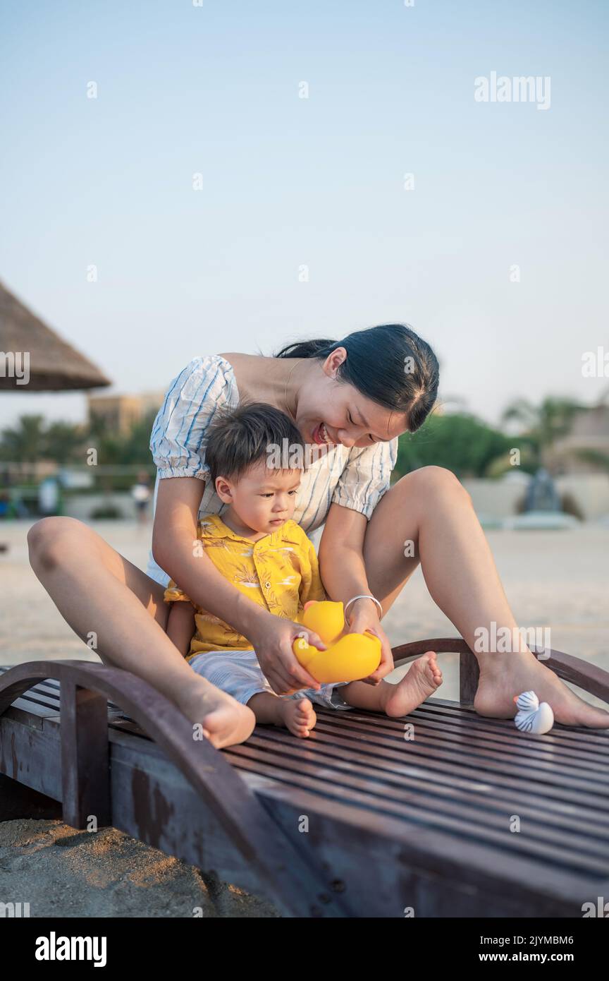 Kleiner Junge auf einem Strandurlaub, der mit seiner Mutter auf der Sonnenliege sitzt und mit der Spielzeug-Ente spielt. Asiatische Frau und ihr ein Jahr alt Kleinkind Junge mit Spaß auf Stockfoto