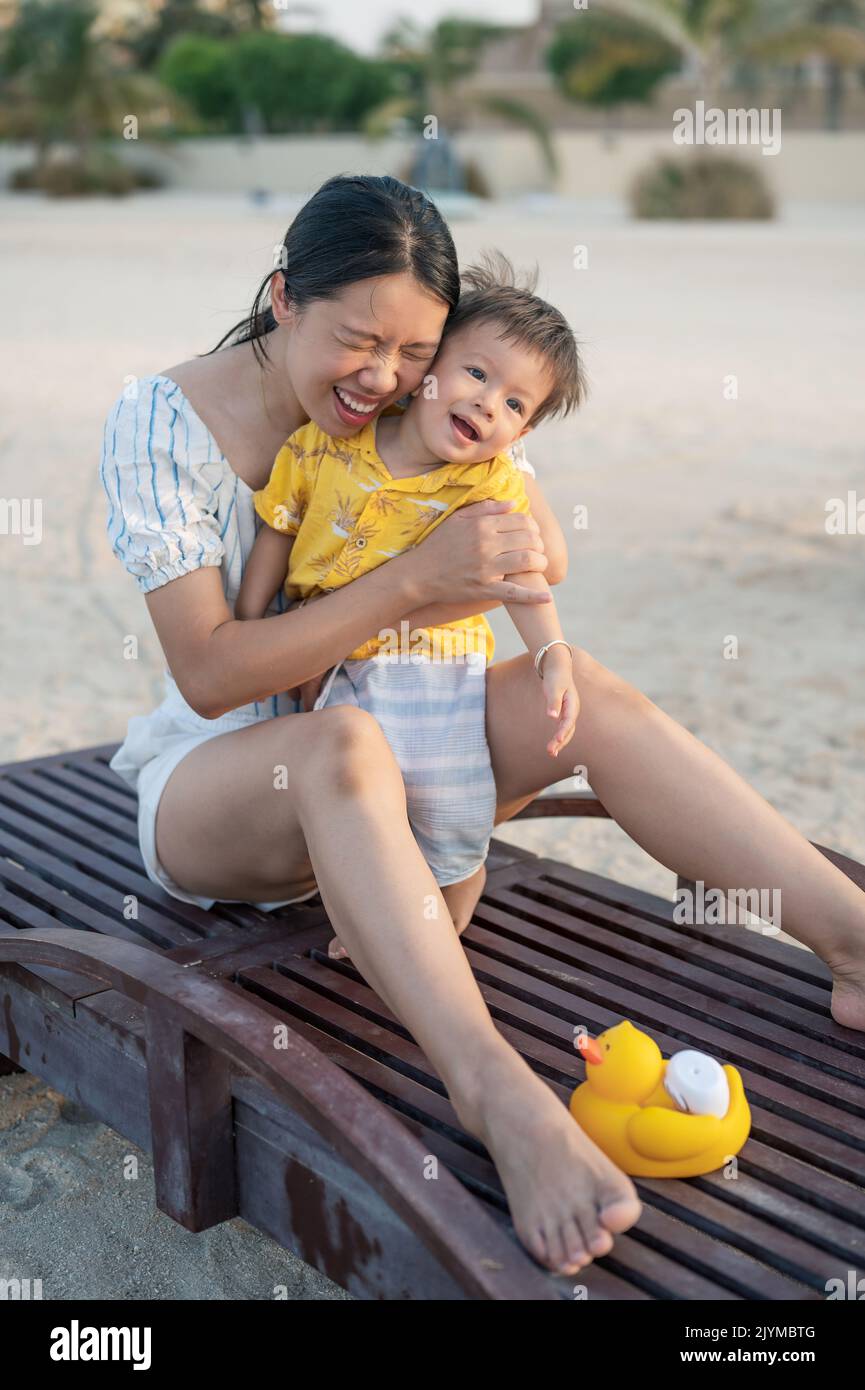 Kleiner Junge auf einem Strandurlaub, der mit seiner Mutter bei Sonnenuntergang auf der Sonnenliege sitzt. Asiatische Frau und ihr einjähriger Säugling Junge mit Spaß im Sommerurlaub Stockfoto