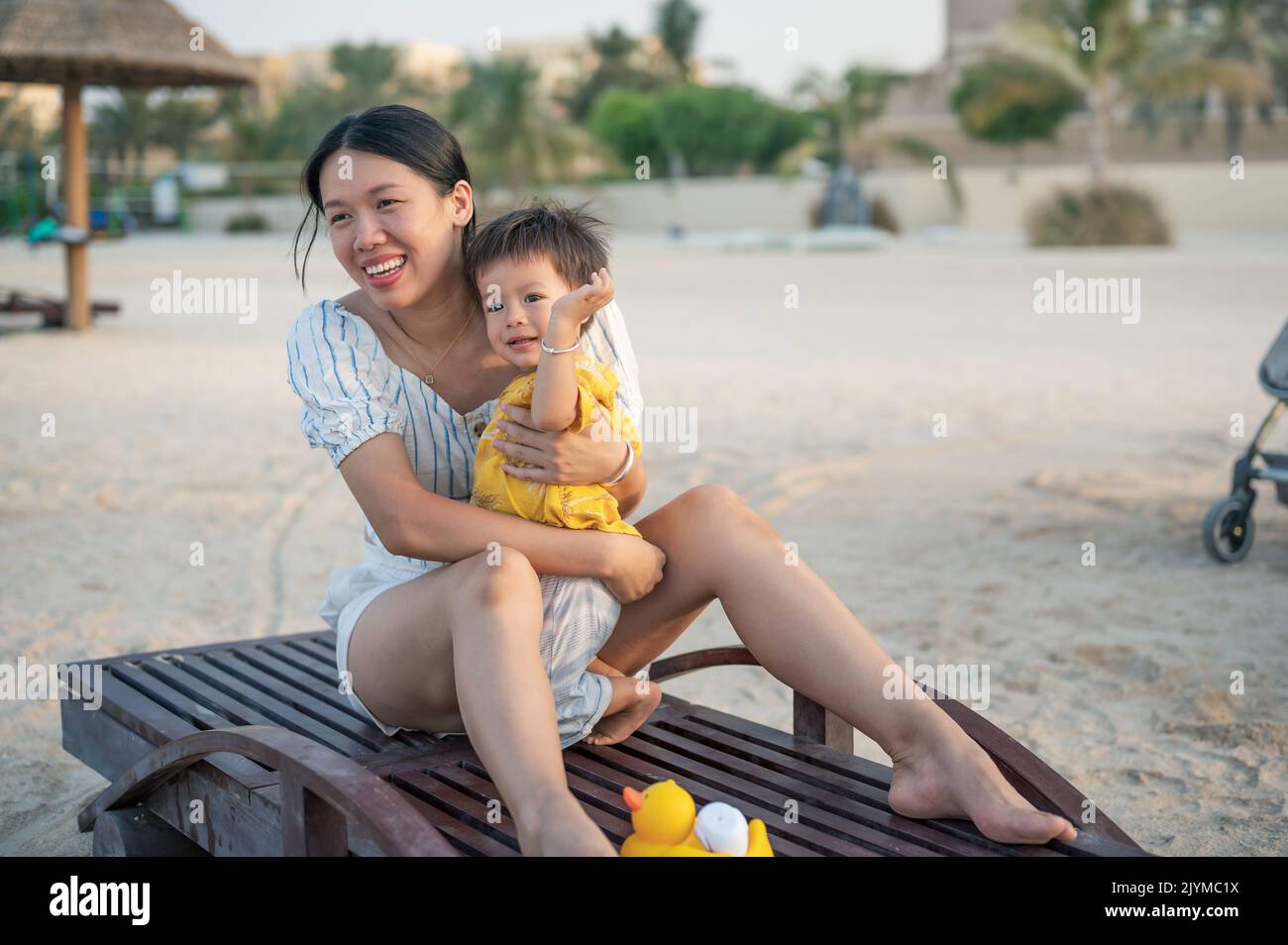 Kleiner Junge auf einem Strandurlaub, der mit seiner Mutter bei Sonnenuntergang auf der Sonnenliege sitzt. Asiatische Frau und ihr einjähriger Säugling Junge mit Spaß im Sommerurlaub Stockfoto