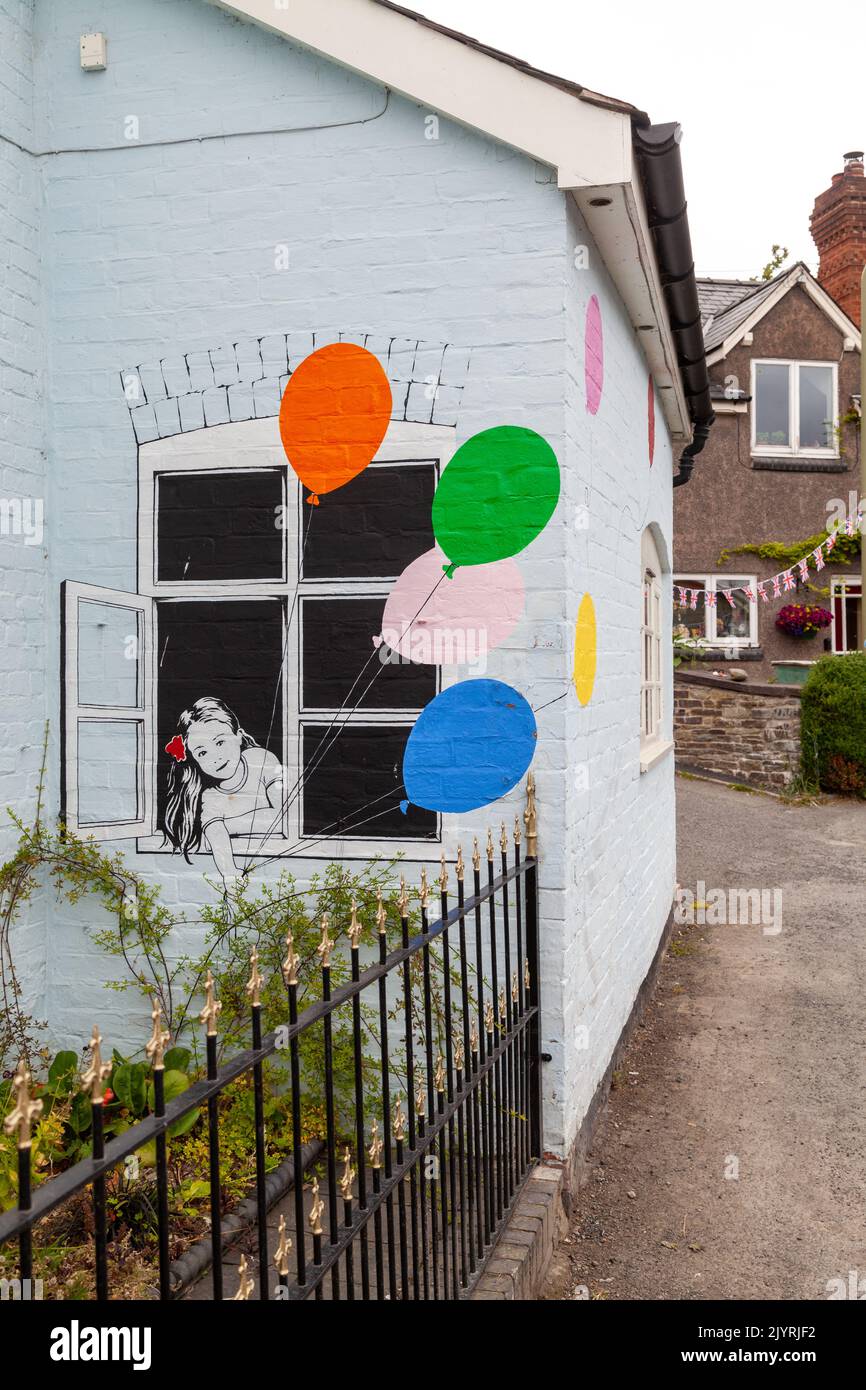 Graffiti-Kunst im Banksy-Stil eines Mädchens mit Luftballons, das aus einem Fenster auf ein Haus im Bishops Castle blickt. Stockfoto