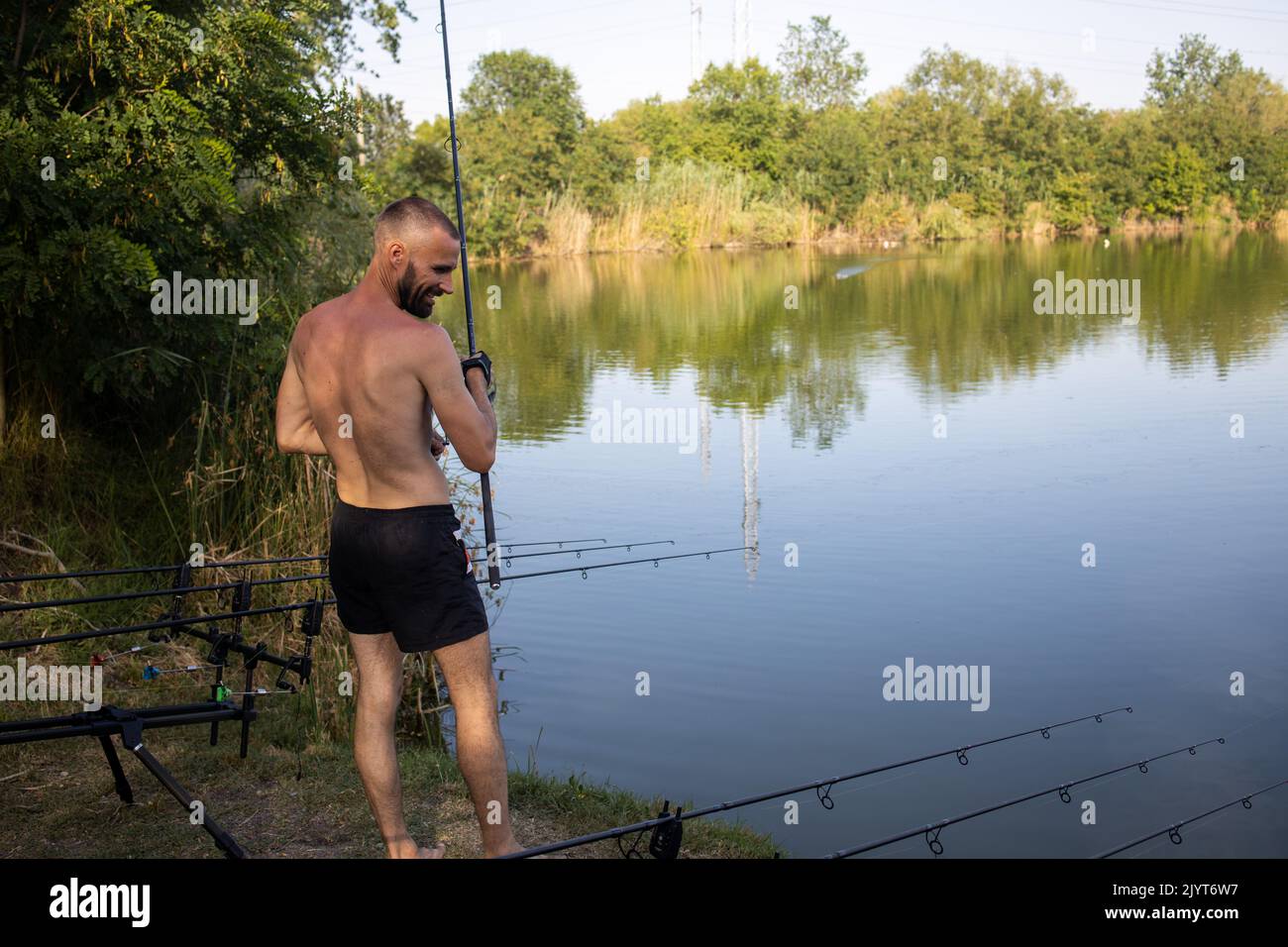 Lächelnder Fischer, der vor dem See ohne Raffen steht, Angelrute hält und andere Angelruten anschaut Stockfoto