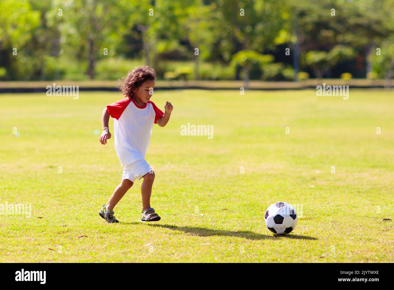 Netter lockiger kleiner Junge, der Fußball spielt. Kinder spielen auf dem Platz im Freien. Entzückende kleine Athlet tritt Ball. Sommerspaß im Freien. Stockfoto