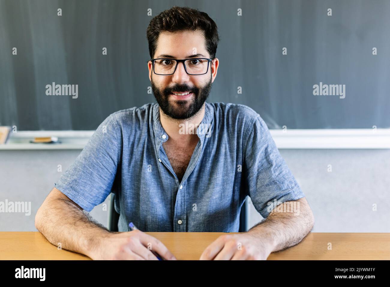 Fröhliches Porträt eines männlichen Lehrers, der im Klassenzimmer am Schreibtisch sitzt Stockfoto