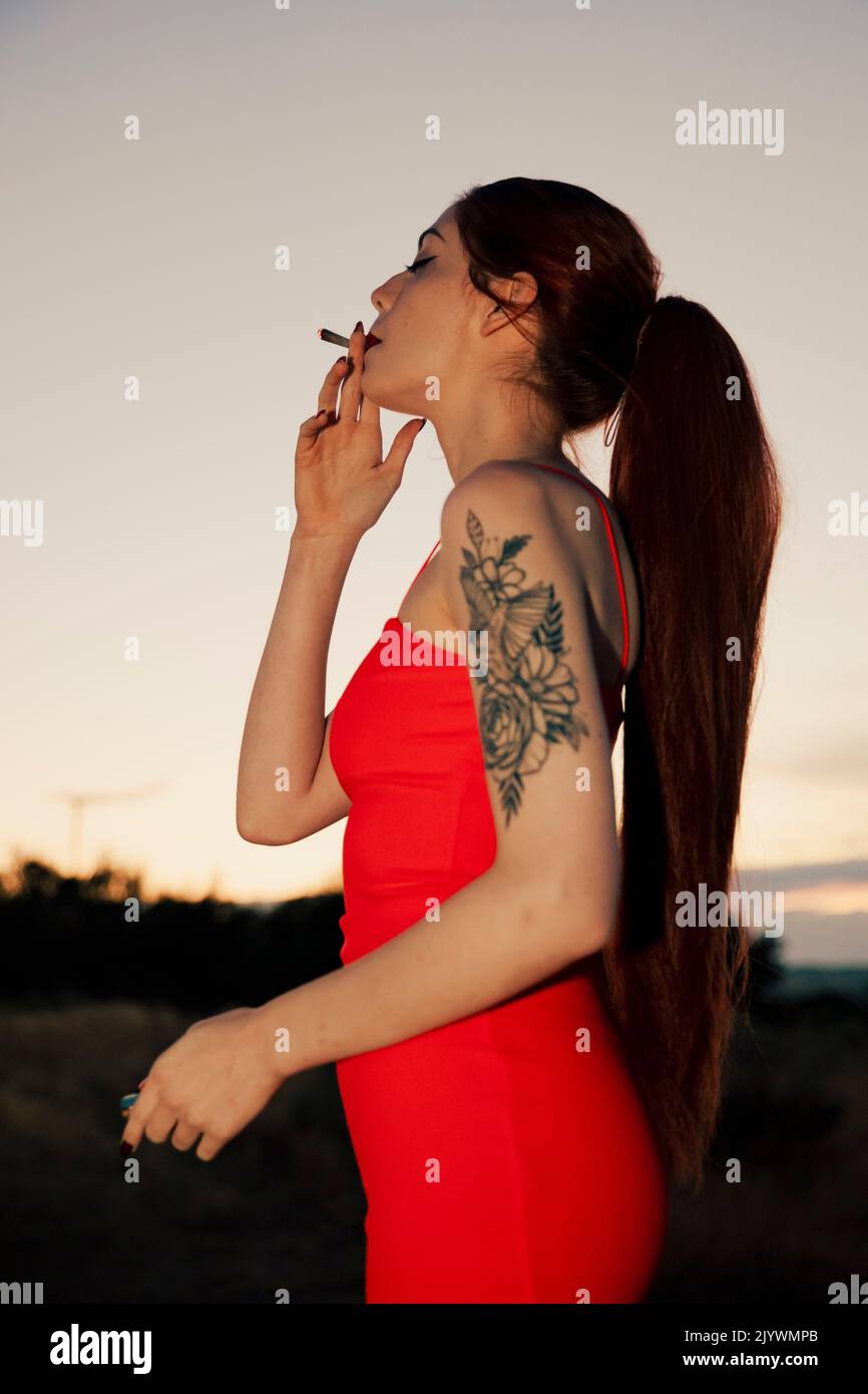 Mädchen in einem roten Kleid, das während des Sonnenuntergangs ein Marihuanagelenk raucht Stockfoto