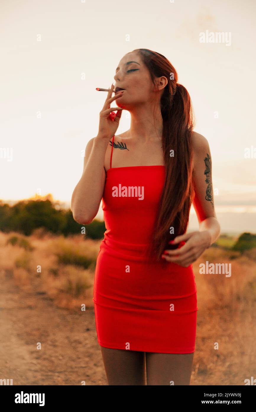 Mädchen in einem roten Kleid, das während des Sonnenuntergangs ein Marihuanagelenk raucht Stockfoto
