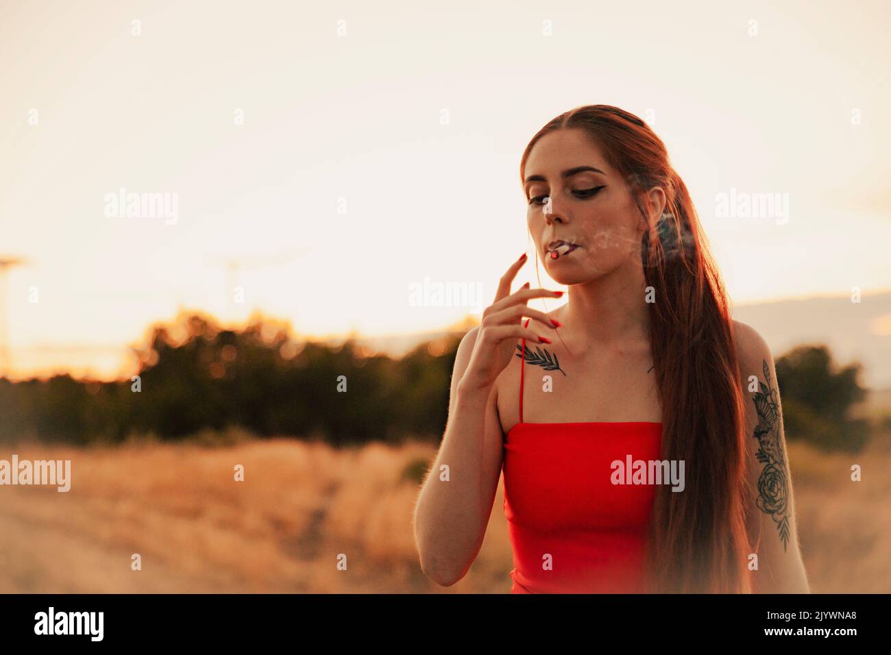 Tätowierte Mädchen in einem roten Kleid rauchen ein Marihuana-Joint während des Sonnenuntergangs Stockfoto