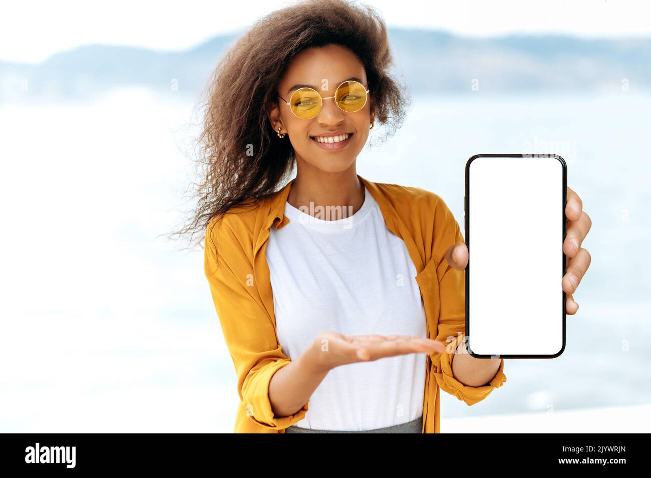 Leerer Raum für Präsentation oder Werbung. Moderne afroamerikanische lockige junge Frau, mit oranger Brille, steht im Freien, zeigt Smartphone mit weißem Mockup-Bildschirm, zeigt Hand, lächelnd Stockfoto